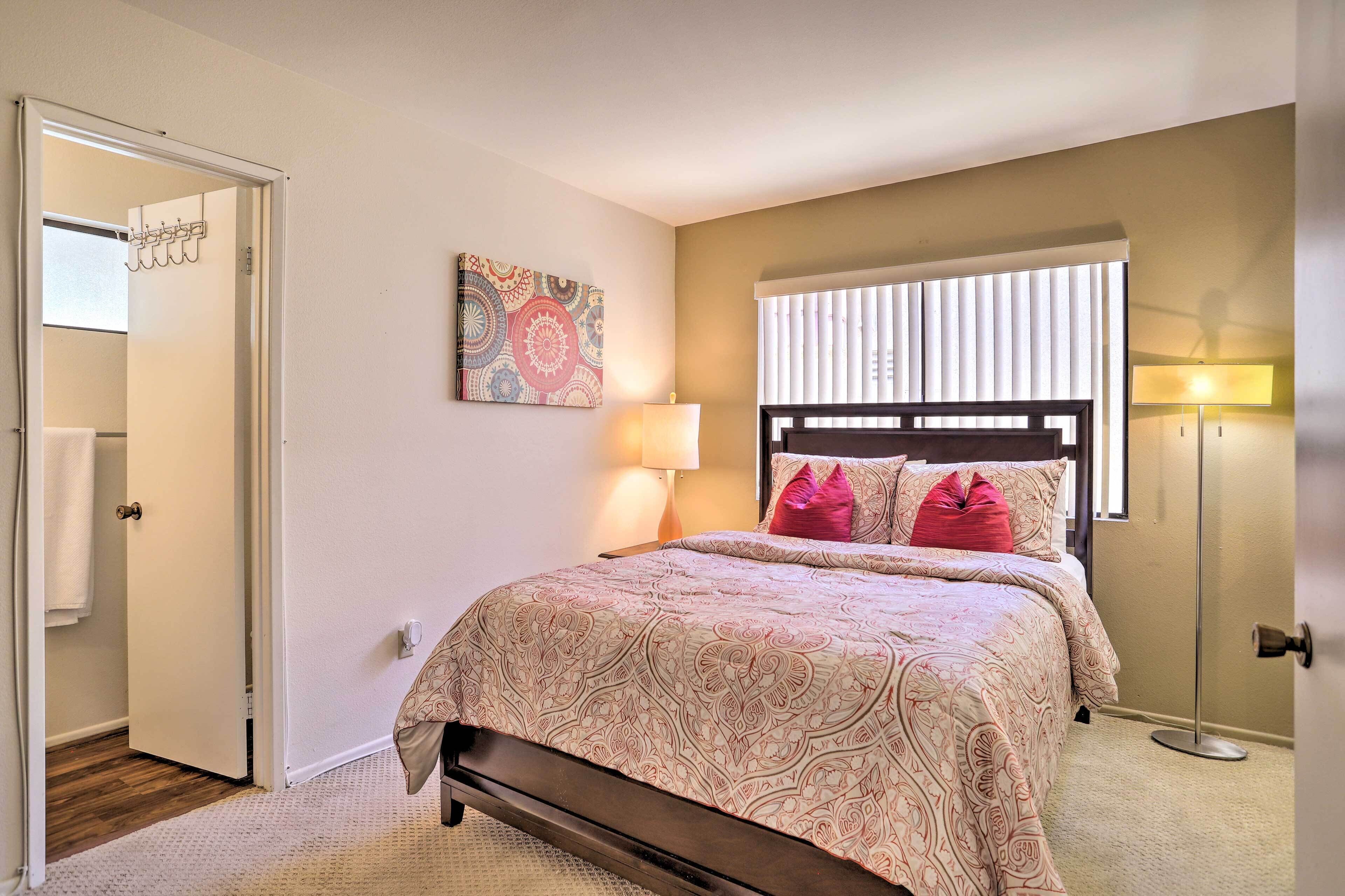 A bedroom with a bed, a bedside lamp, a window with blinds, and a door leading to another room.