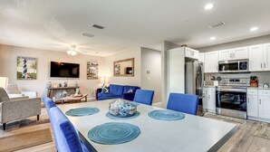 A modern kitchen with white cabinets, stainless steel appliances, and a dining area with blue chairs and a large table. - Colorful Cottage 1 Mi to Historic District! (St. Augustine)