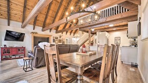 Un salon spacieux avec une table à manger en bois, des chaises et un grand canapé. Un téléviseur est fixé au mur, et un escalier mène à un niveau supérieur.