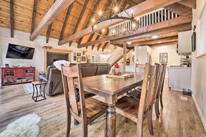 Ein geräumiges Wohnzimmer mit einem Holzessttisch, Stühlen und einem großen Sofa. Ein Fernseher ist an der Wand befestigt, und eine Treppe führt zu einer oberen Ebene.