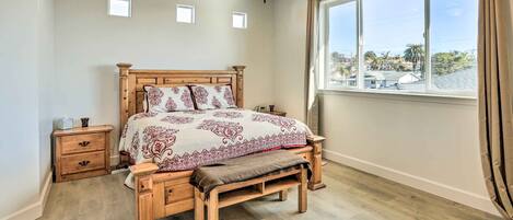 A bedroom with a wooden bed, a nightstand, a bench, a ceiling fan, and a large window with a view of palm trees and buildings.
