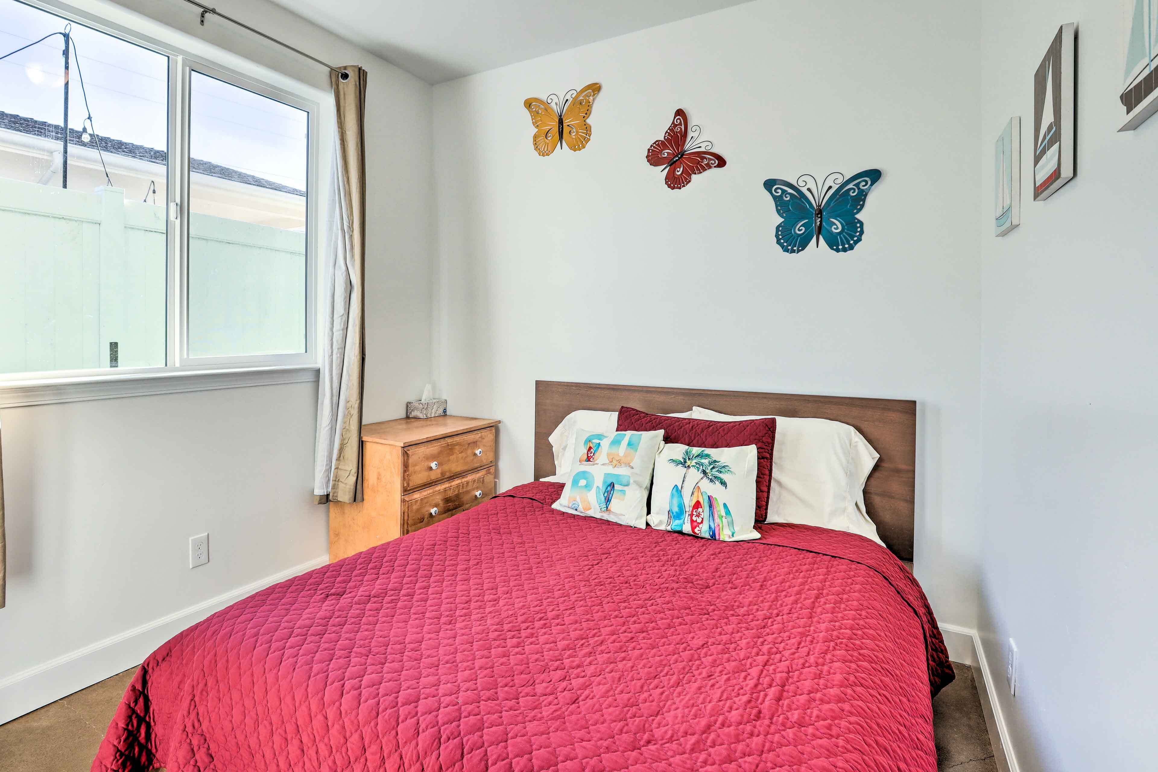 A bedroom with a red quilted bedspread, a wooden nightstand, a dresser, and a window with curtains. There are three butterfly wall decorations.
