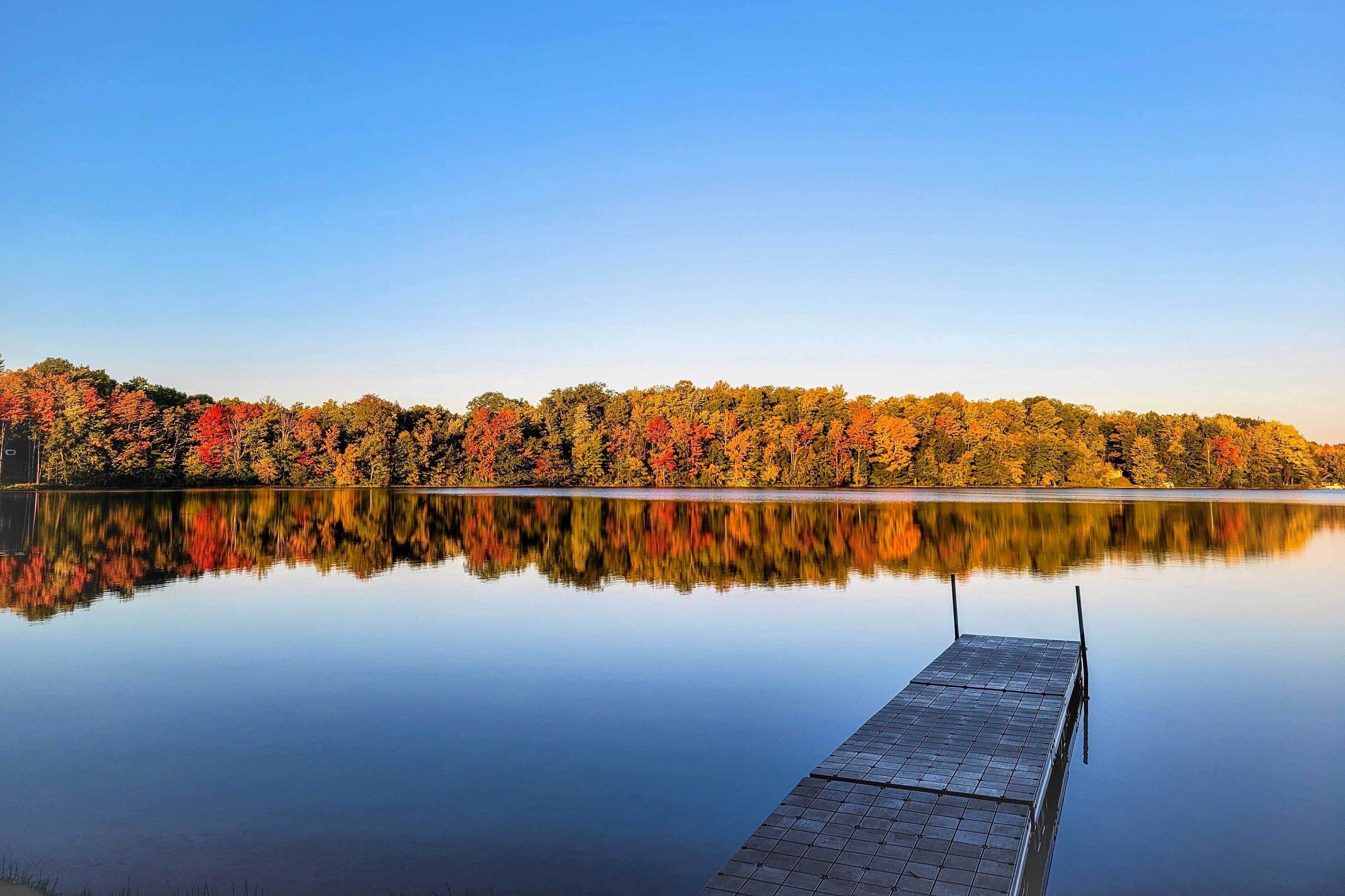 A calm lake with a dock extending into the water, surrounded by trees with autumn foliage.