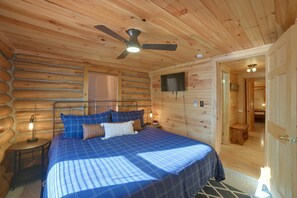 Un dormitorio de cabaña con una cama, un ventilador de techo, un televisor montado en la pared y una puerta de madera que da a otra habitación.