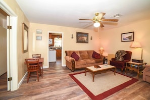 A living room with a wooden floor, a ceiling fan, a sofa, a coffee table, a dining table with chairs, and a kitchen in the background. - Fredericksburg Home 8 Blocks to Main St + Wineries (Fredericksburg)