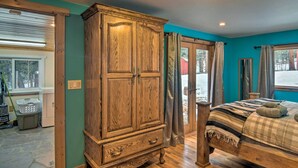 A bedroom with a wooden wardrobe, a bed with a checkered blanket, and a view of the outdoors through a glass door. - Allenspark Vacation Rental Near Estes Park! (Allenspark)