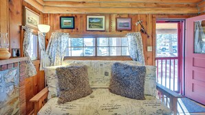 Ein gemütliches Cabin-Zimmer mit Holzbalkendecke, einem Bett mit gemusterten Textilien, einem Fenster mit Vorhängen und einer rosafarbenen Tür.