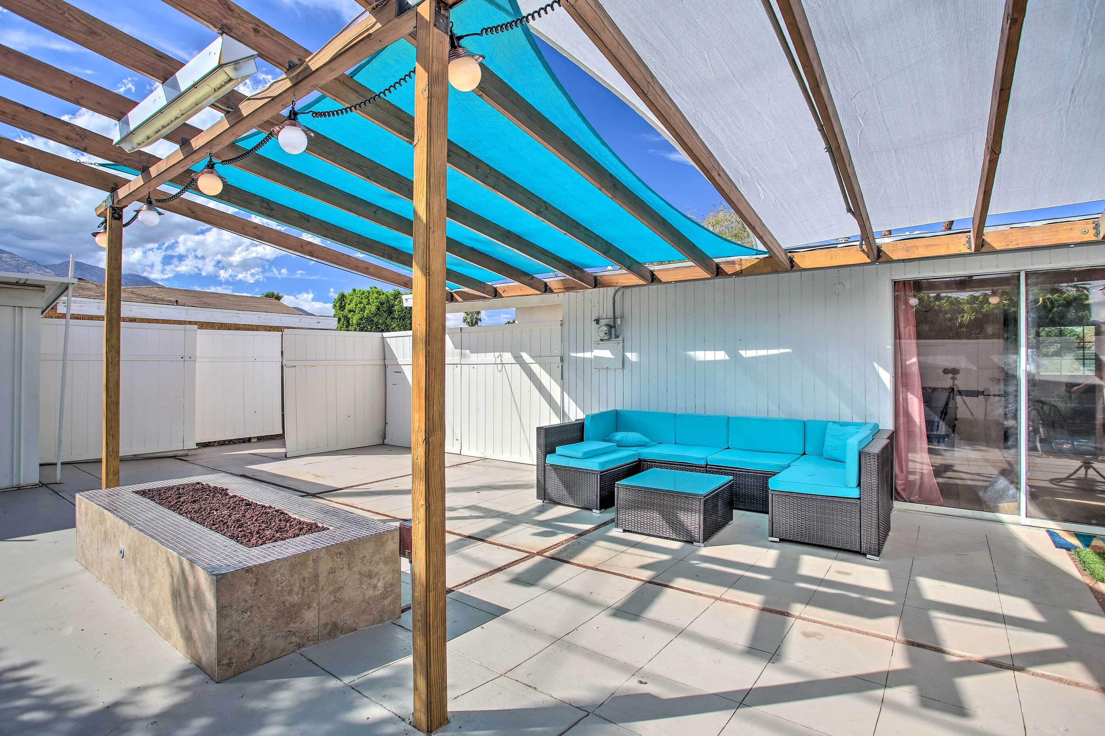 Un patio exterior con un sofá azul, un pozo de fuego y una pérgola con luces colgantes.