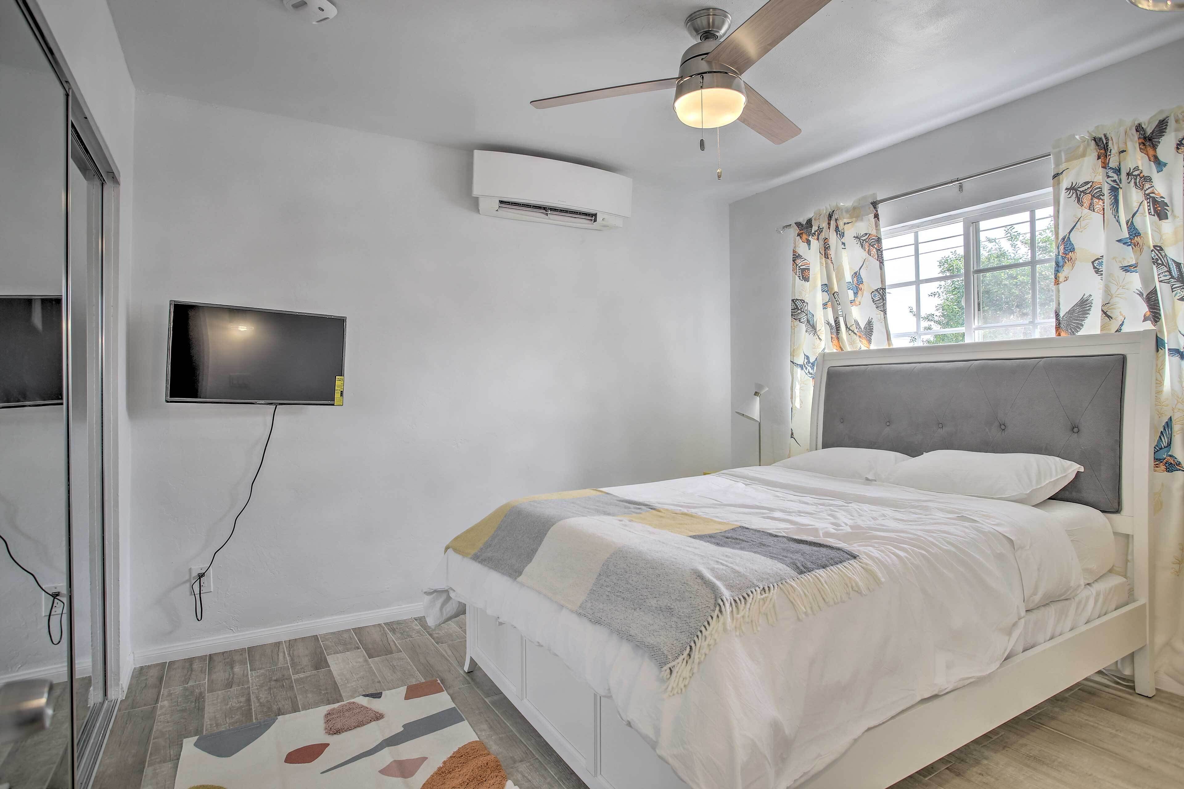 A bedroom with a bed, ceiling fan, air conditioning unit, TV, and a window with curtains.