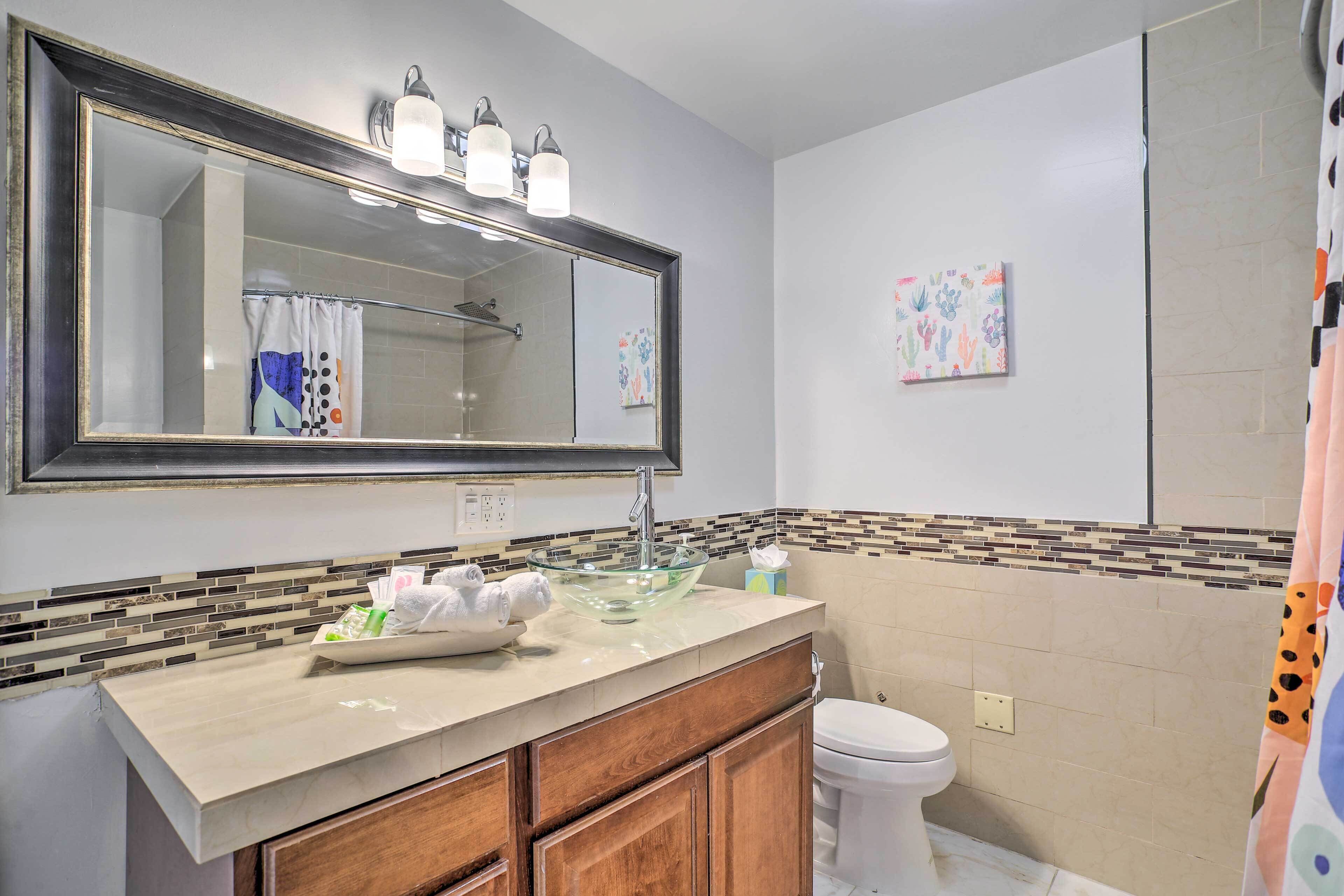 A bathroom with a large mirror, a glass sink, a toilet, and a shower area.