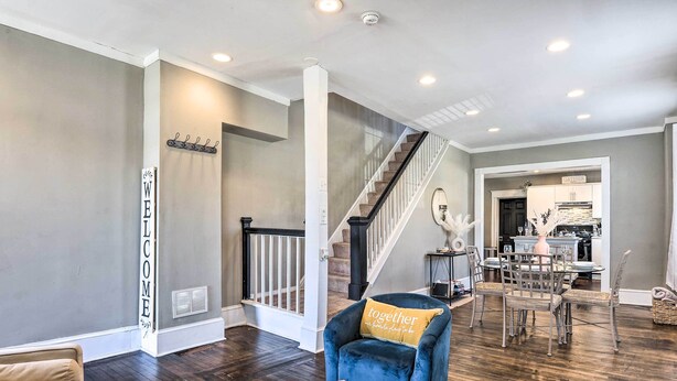 A spacious living room with a dark wood floor, a blue armchair with a 'together' cushion, a beige sofa, and a coffee table with a woven centerpiece. A staircase leads to a dining area with a chandelier. - Baltimore Vacation Rental Near Dtwn Towson! (Baltimore)