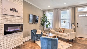 A living room with a brick fireplace, a flat-screen TV, a blue armchair, a white sofa, and a large plant. - Baltimore Vacation Rental Near Dtwn Towson! (Baltimore)