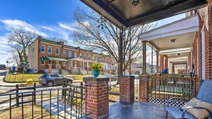 A covered porch with a bench and a potted plant, overlooking a street with parked cars and a row of brick townhouses. - Baltimore Vacation Rental Near Dtwn Towson! (Baltimore)