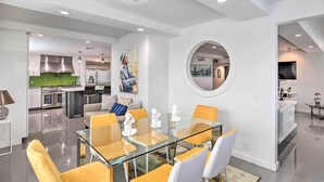 A modern kitchen with stainless steel appliances, a dining area with a glass table and yellow chairs, and a living space with a round mirror and artwork.