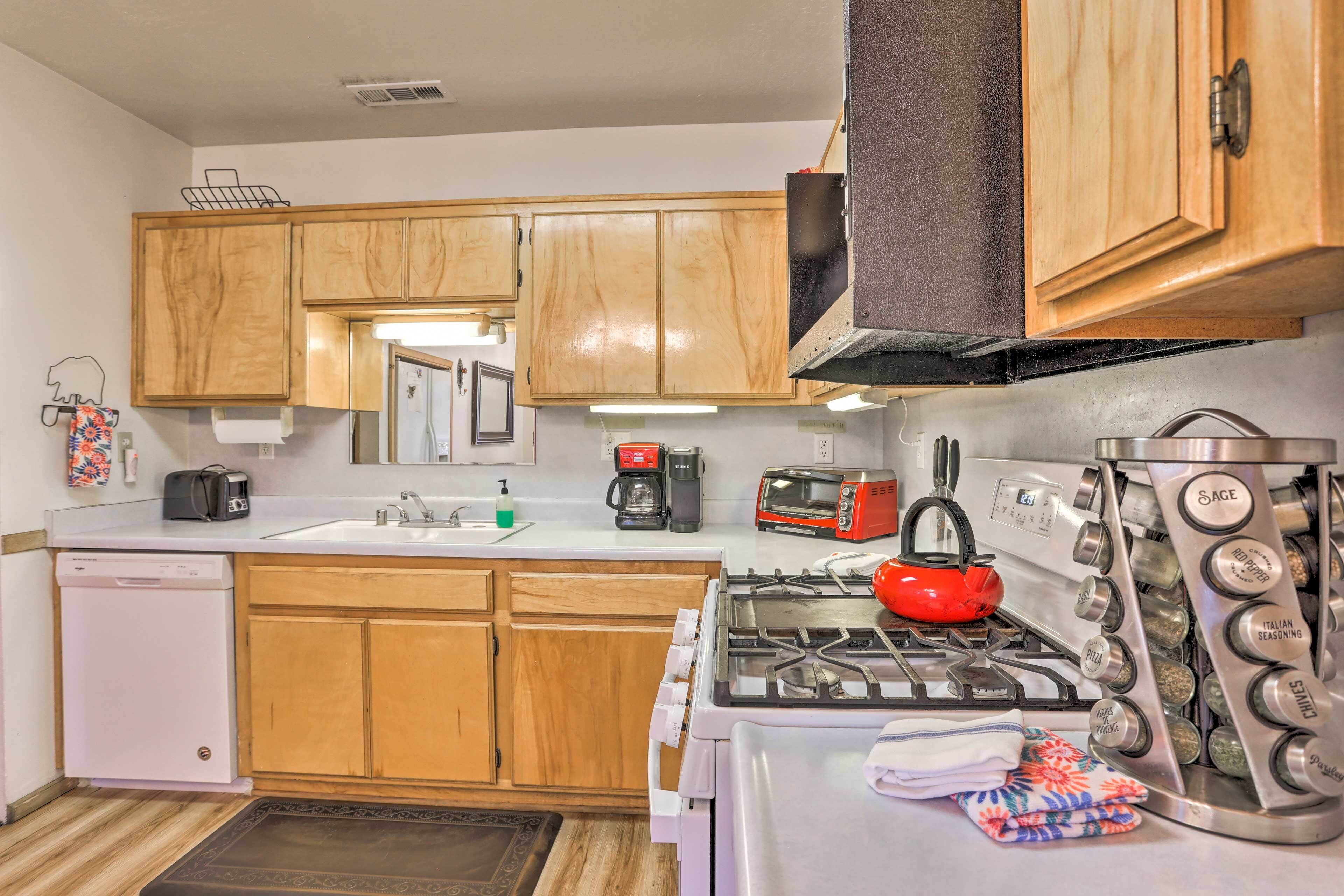 A compact kitchen with wooden cabinets, a white dishwasher, a red kettle, and a stainless steel stove with a spice rack.