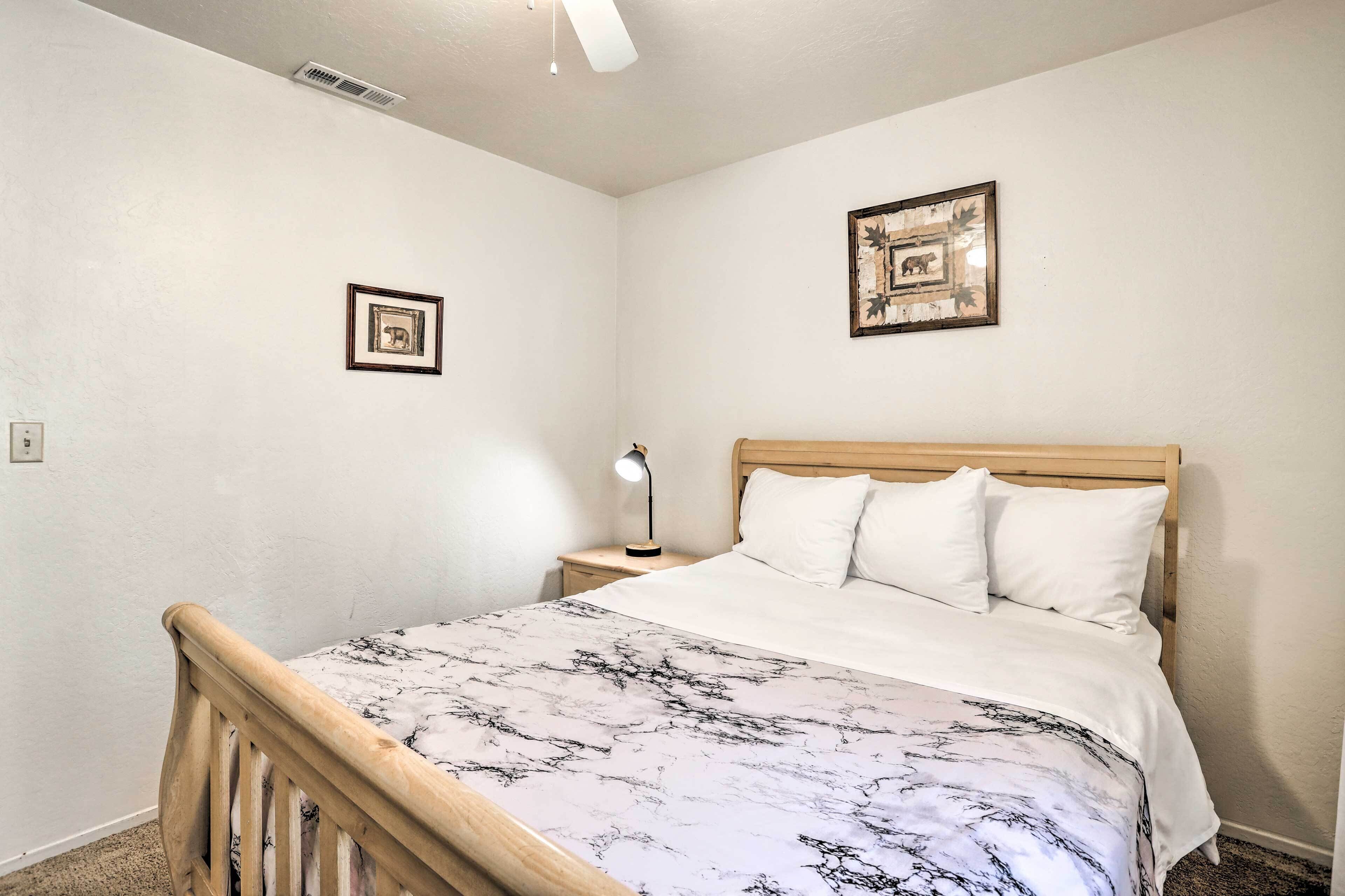A bedroom with a wooden bed frame, white bedding, a nightstand with a lamp, and two framed pictures on the walls.