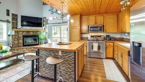 Une cuisine moderne avec des meubles en bois, une cheminée en pierre, un grand îlot central avec des tabourets et des appareils électroménagers en acier inoxydable.
