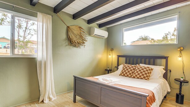 A bedroom with a bed, wooden floor, a window with curtains, an air conditioner, and a small table with a lamp. - Group Friendly! Gem 5 Mi to Joshua Tree Nat'l Park (Joshua Tree)