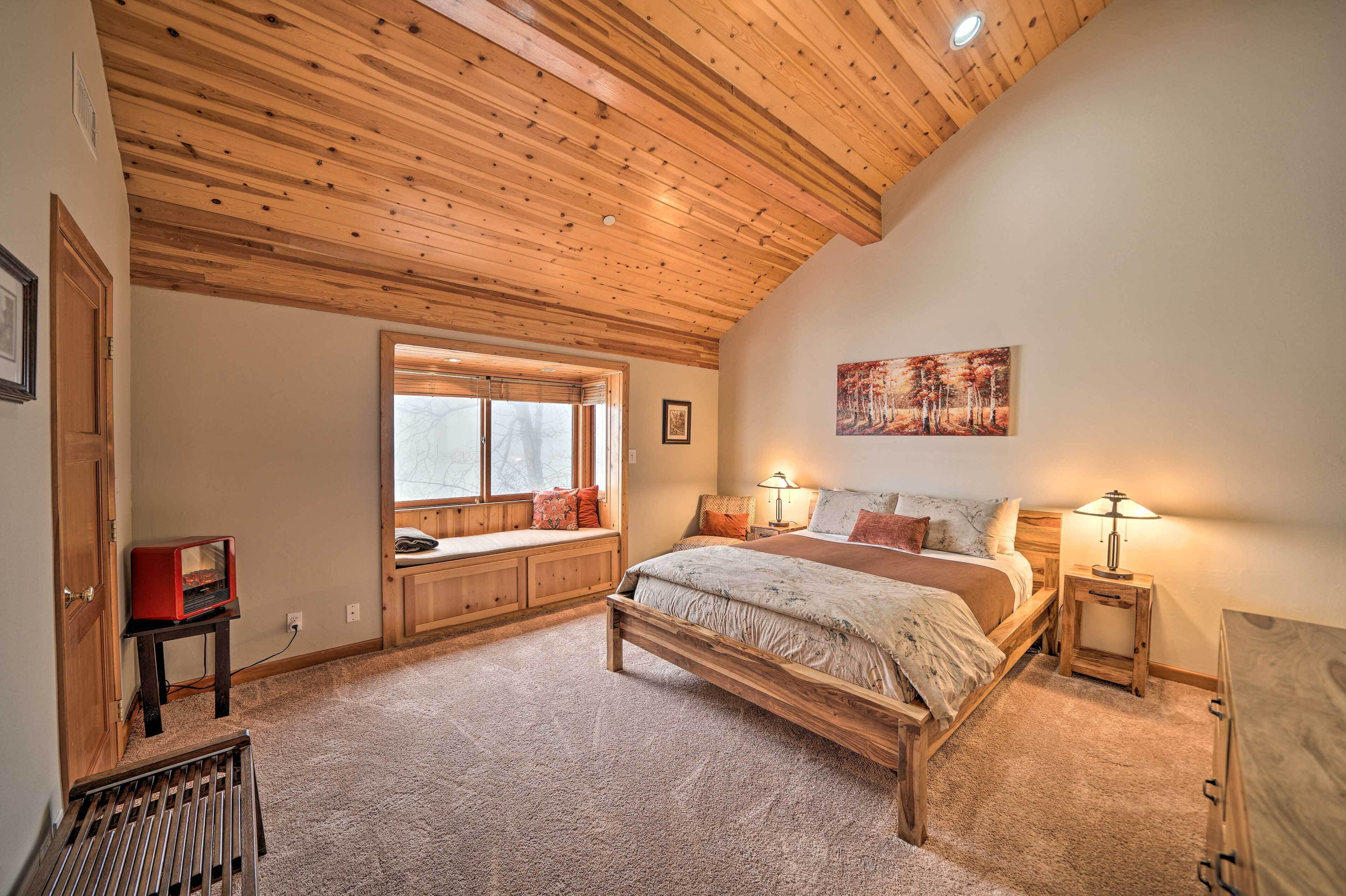 A bedroom with a wooden ceiling, a bed with a headboard, a window with blinds, a bedside table with a lamp, and a carpeted floor.
