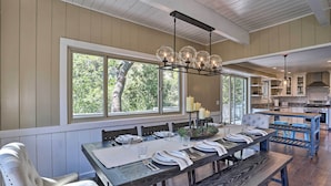 A dining area with a long wooden table set for six, a chandelier, and large windows with a view of trees.