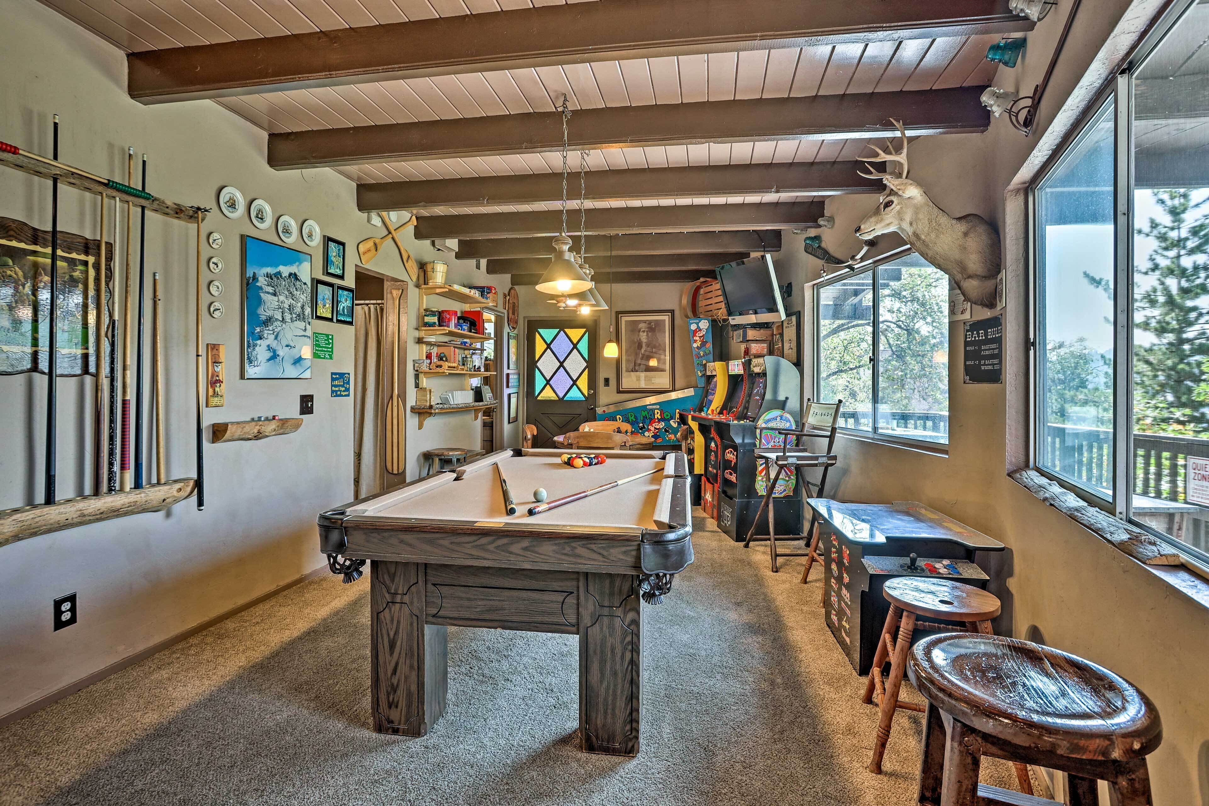 A pool table in a room with wooden beams, a deer head mounted on the wall, and a bar area with arcade games.