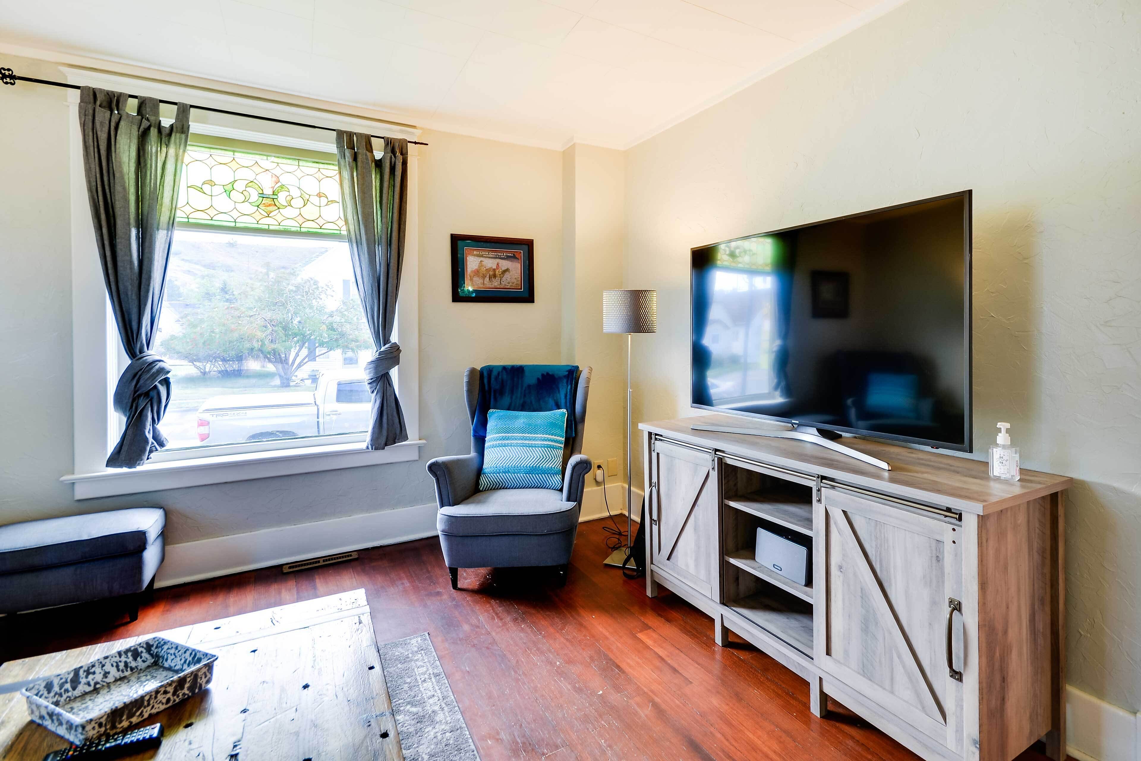 A living room with a large flat-screen TV, a comfortable armchair, a sofa, a wooden cabinet, and a window with curtains.