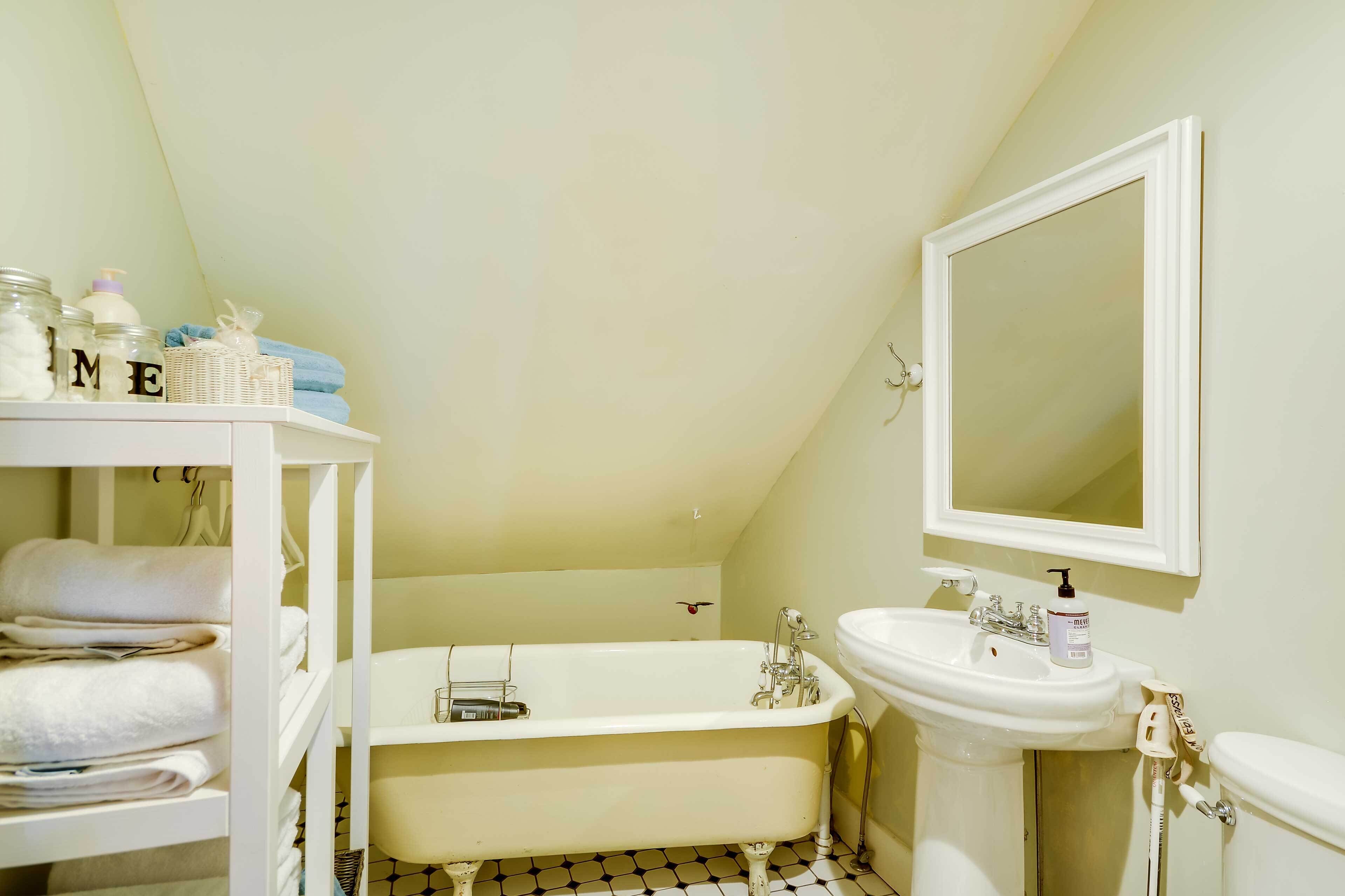 A bathroom with a bathtub, a sink, and a mirror. There are towels stacked on shelves and a basket with toiletries.