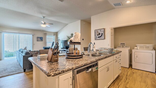 A modern kitchen with granite countertops, stainless steel appliances, and a dishwasher. There is a dining area with a gray sofa and a dining table. - Golfer's Paradise - The Villages in Wildwood! (Wildwood)