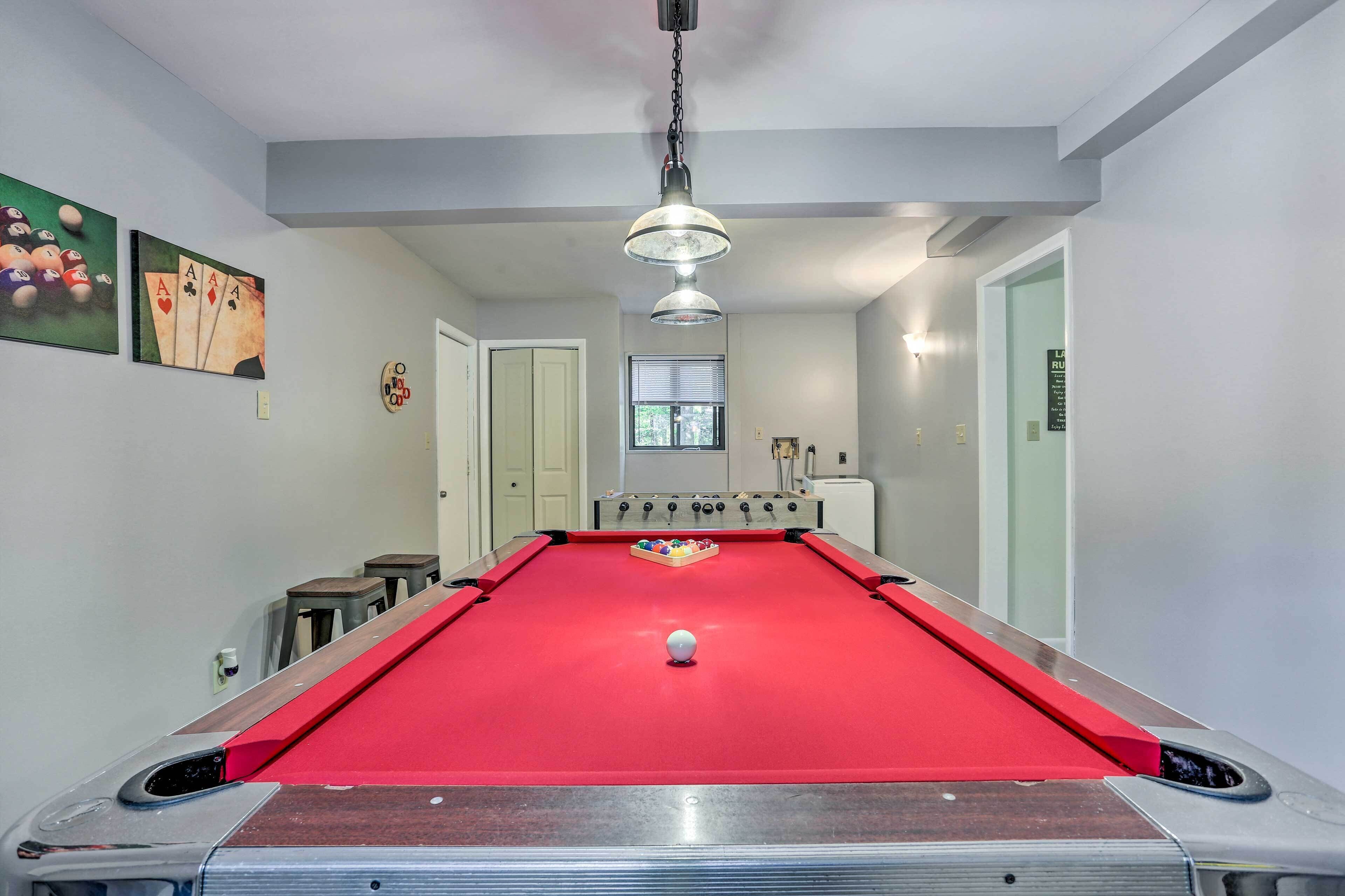 A pool table with a red felt surface, a white cue ball, and a foosball table in the background.