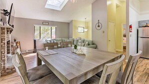 Un coin repas avec une grande table en bois, des chaises et une cheminée en briques. On aperçoit un canapé et une fenêtre au fond.