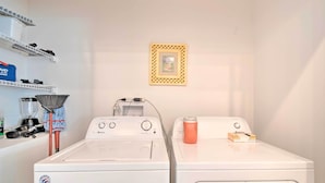 Un área de lavandería con dos lavadoras de carga frontal, un estante con productos de limpieza y un cuadro en la pared.
