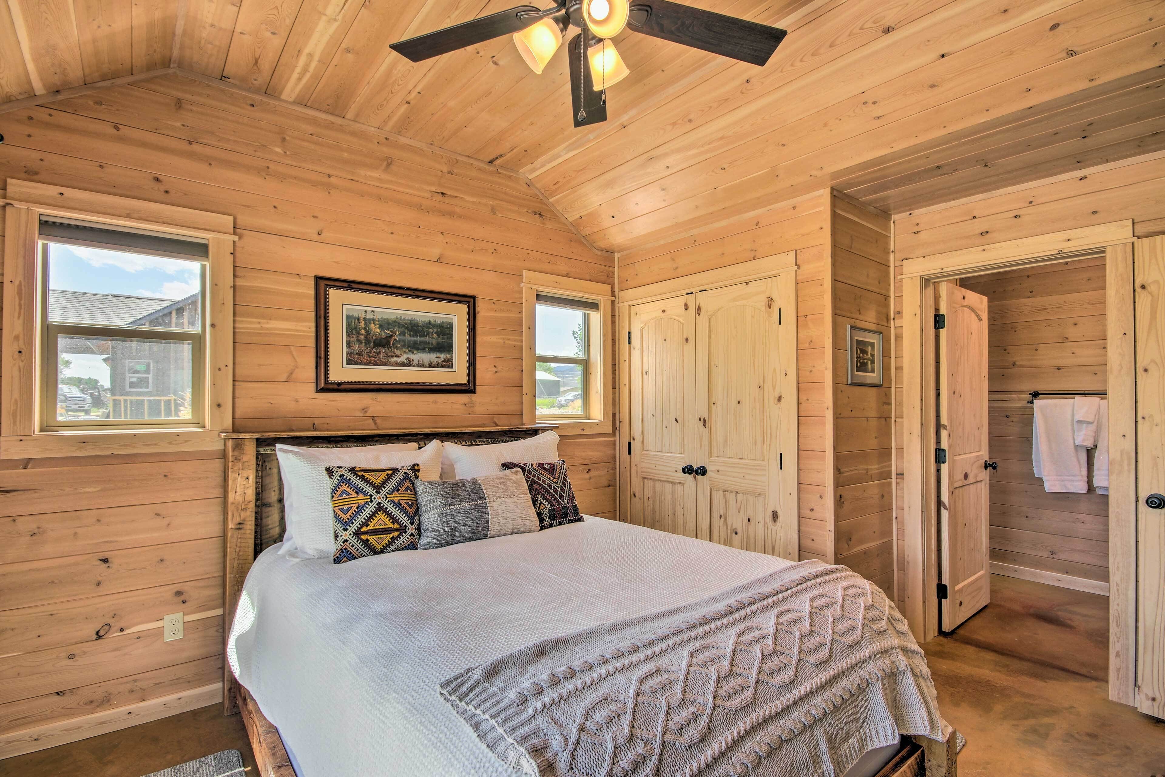 Une chambre à coucher dans une cabane en bois, avec un lit, des oreillers, un ventilateur de plafond et une porte donnant accès à une salle de bain.