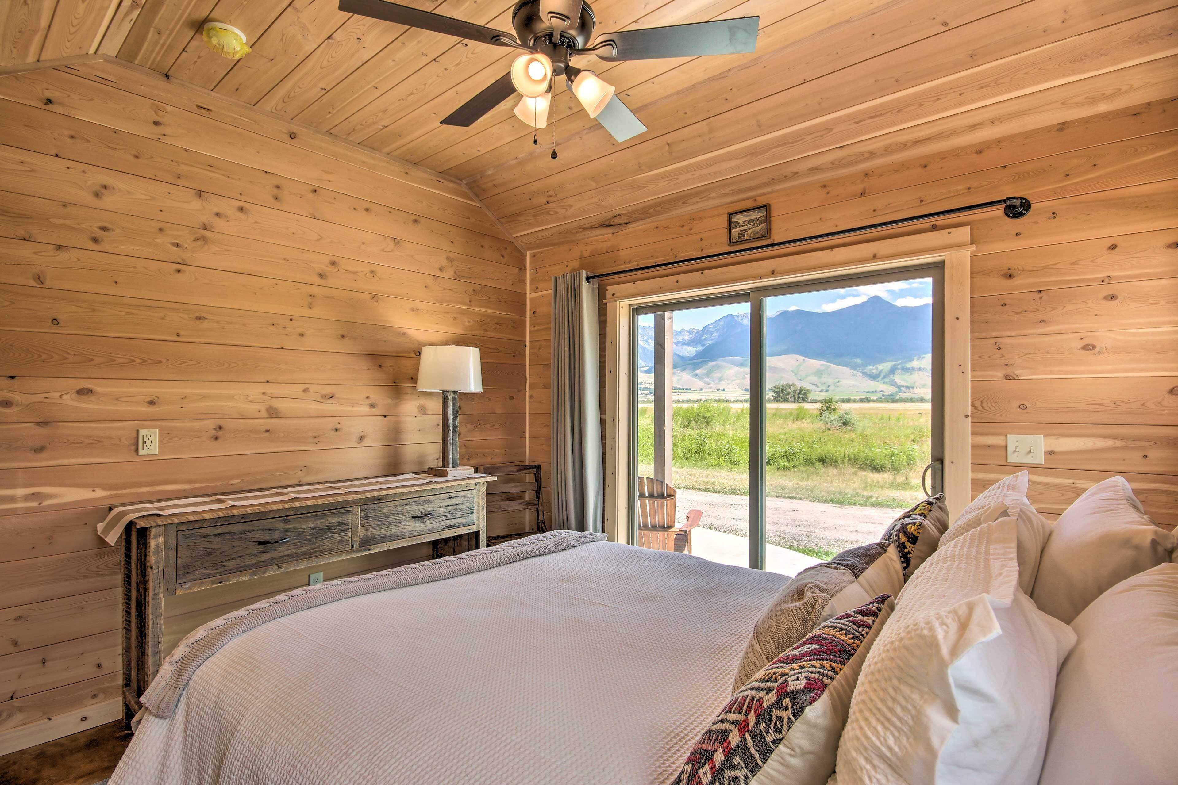 A wooden cabin bedroom with a bed, a ceiling fan, a window with a mountain view, and a rustic dresser.