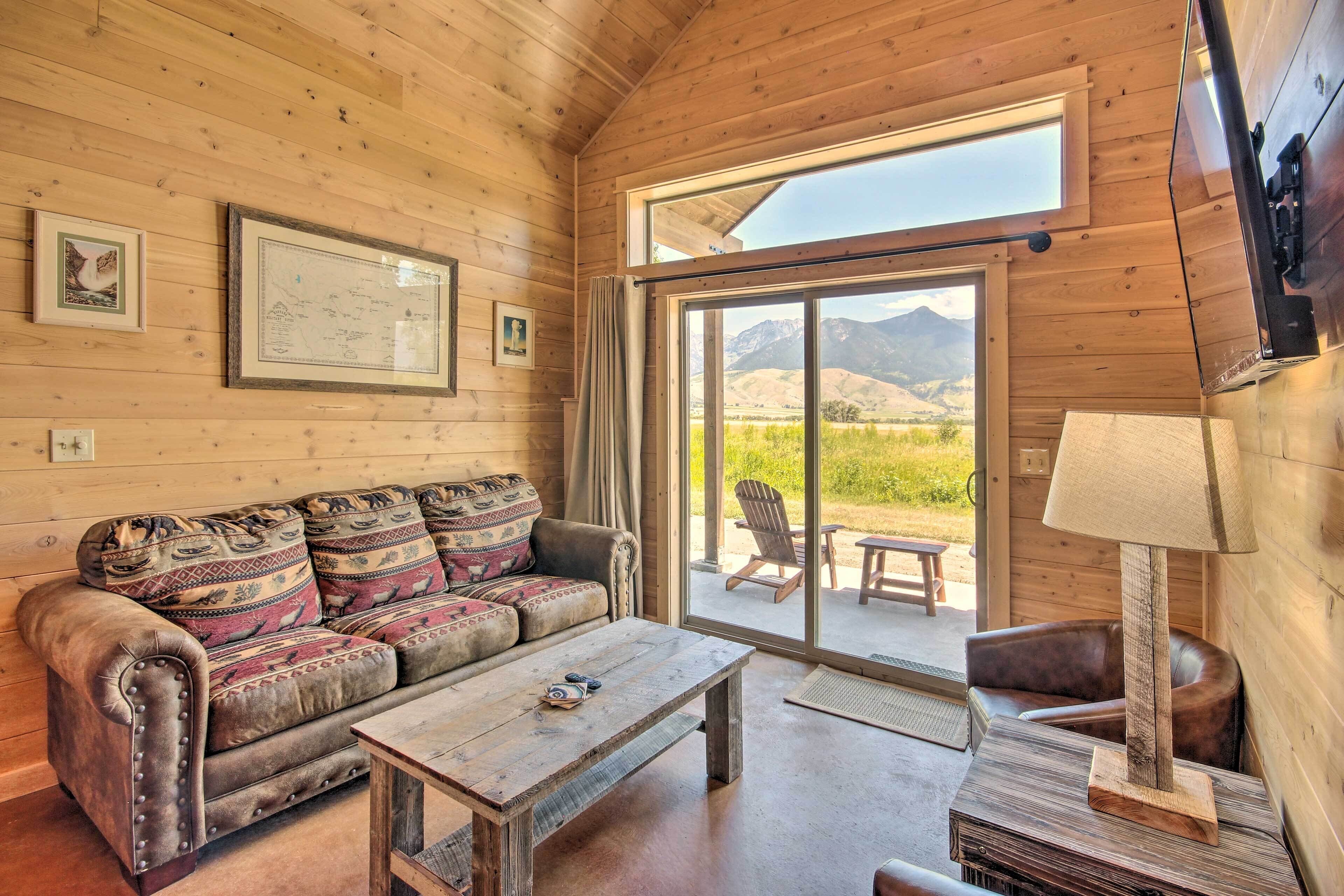 A cozy cabin interior with a wooden ceiling, a sofa with patterned cushions, a wooden coffee table, a lamp, and a sliding glass door leading to an outdoor patio with a chair and a table.
