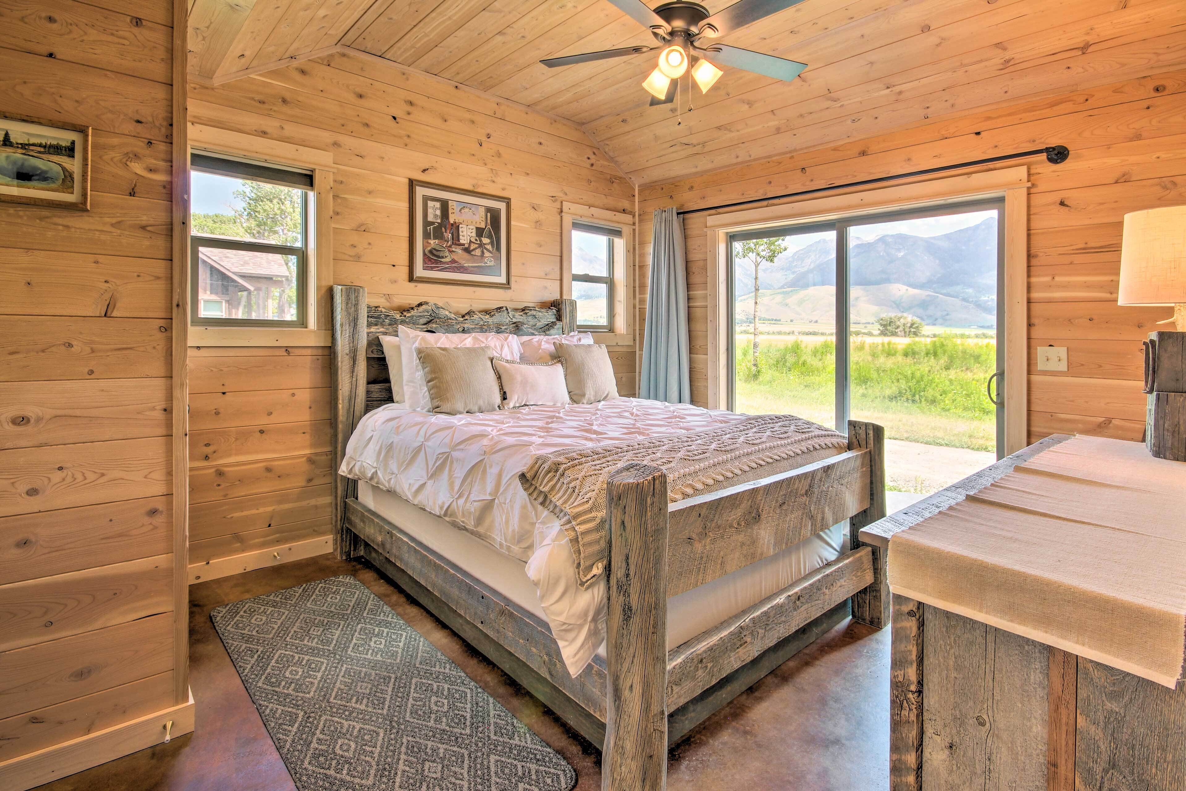 A wooden cabin bedroom with a bed, a nightstand, a ceiling fan, and large windows offering a view of the outdoors.