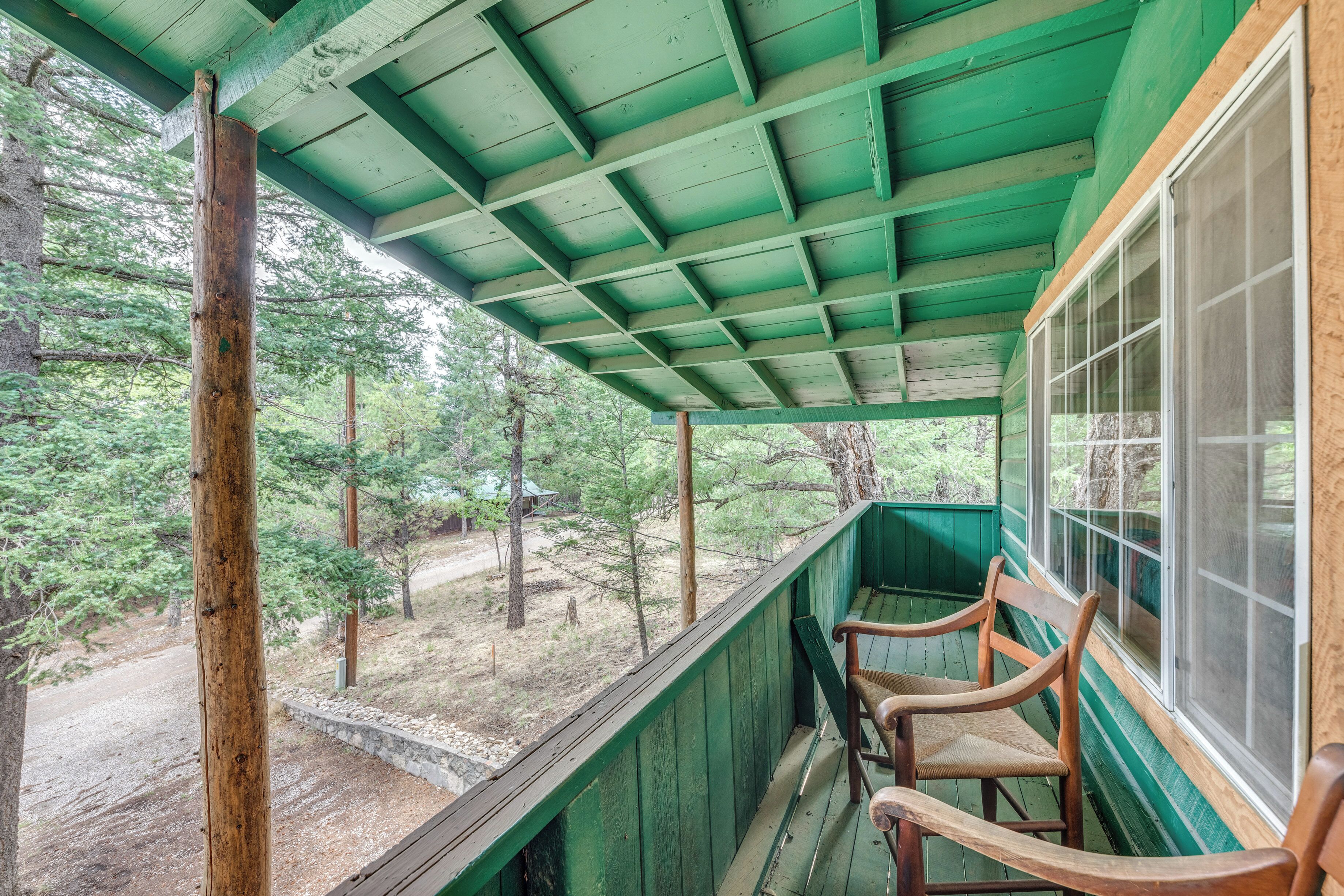 Eine überdachte Veranda mit Holzstühlen und Blick auf eine bewaldete Landschaft, sowie ein Holzkonstrukt mit grüner Dachdeckung.