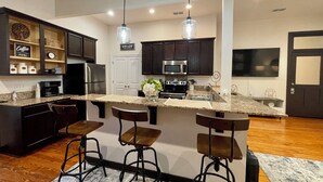 Una cocina moderna con armarios oscuros, electrodomésticos de acero inoxidable y una encimera de mármol. Hay tres taburetes de bar y lámparas colgantes sobre la encimera.