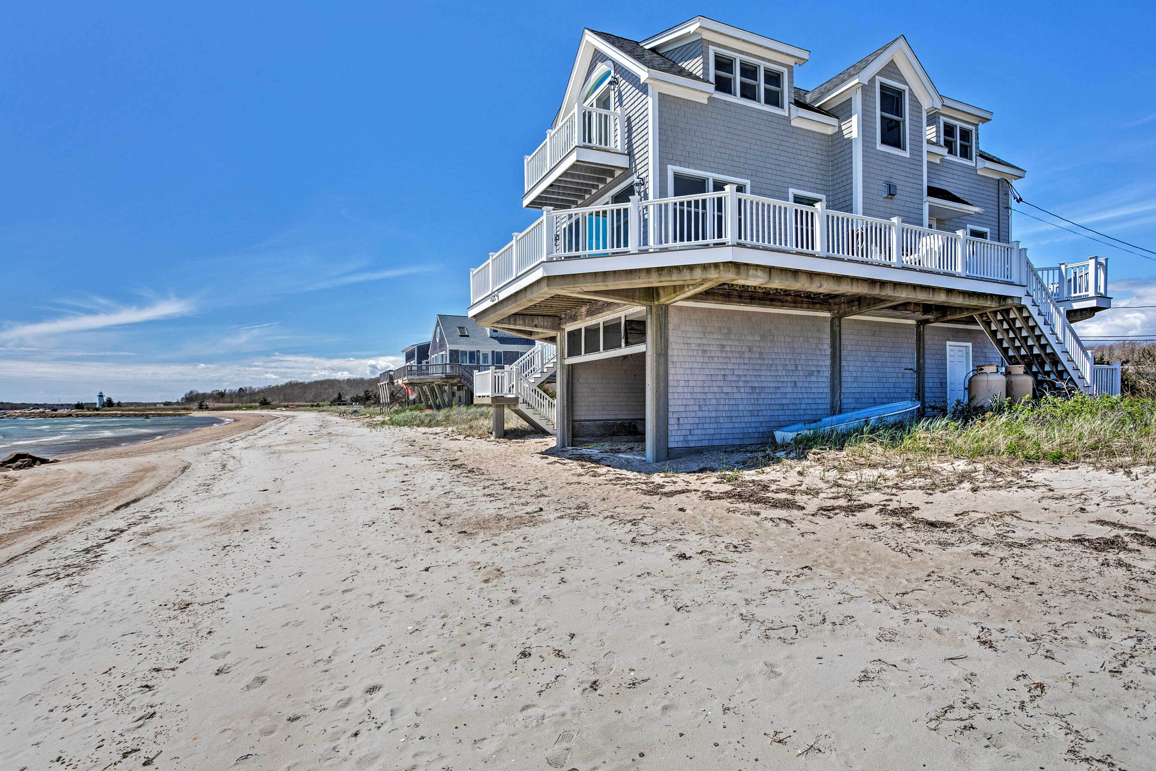 A beachfront house with a balcony, a staircase leading to the upper floor, and a sandy beach in the foreground.