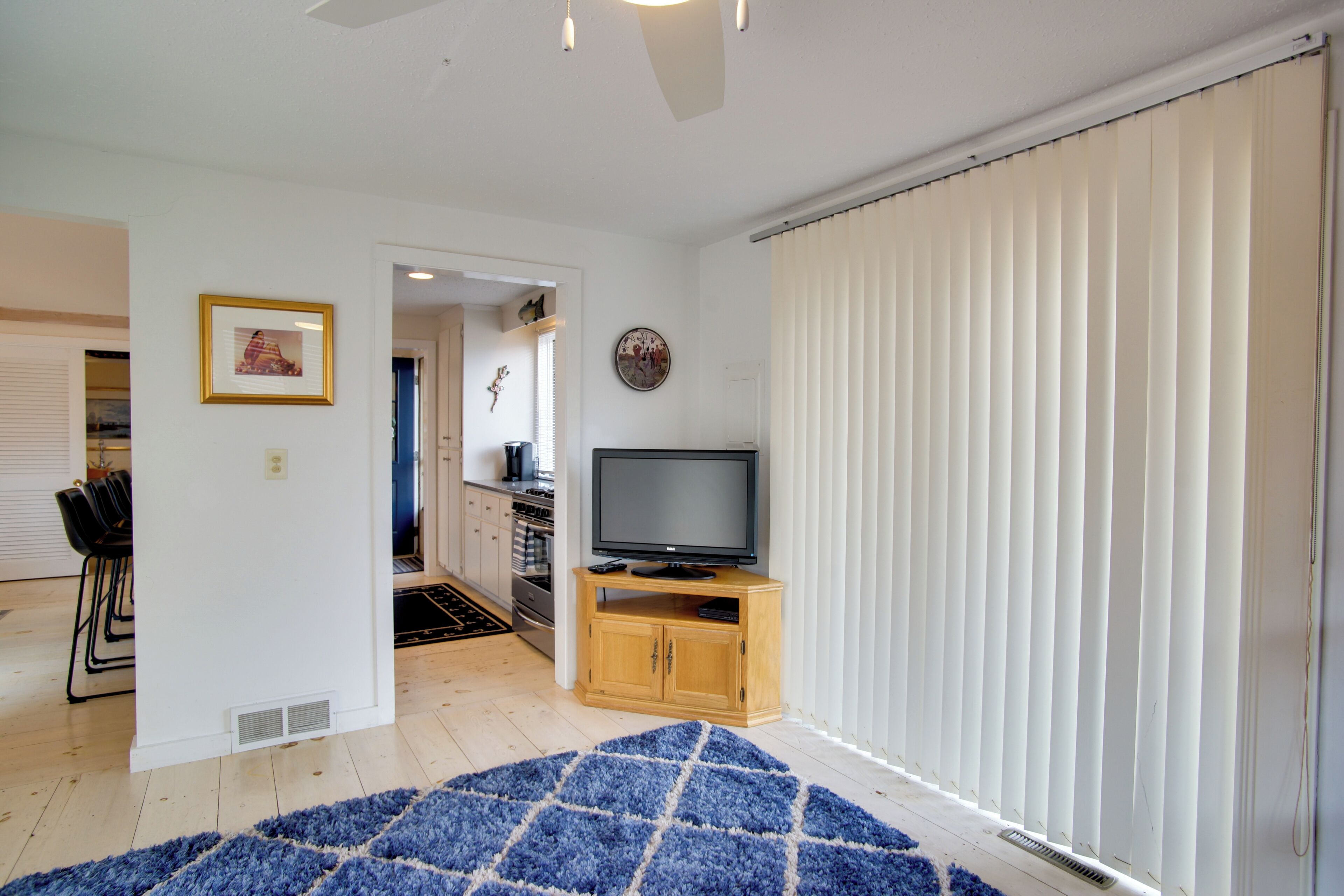 A living room with a TV on a wooden stand, a blue rug, and a sliding glass door with blinds.