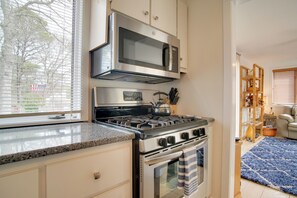 A kitchen with a gas stove, stainless steel microwave, and oven. There is a window with blinds, a blue rug, and a wooden shelf with items. - Charming Cape Cod Home w/ Patio, 2 Mi to Beach! (Mashpee)