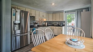 A compact kitchen with white cabinets, a stainless steel refrigerator, a wooden dining table, and a view through the window. - Walk to Beach: Quaint Dennis Home w/ Deck + Grill! (Dennis Port)
