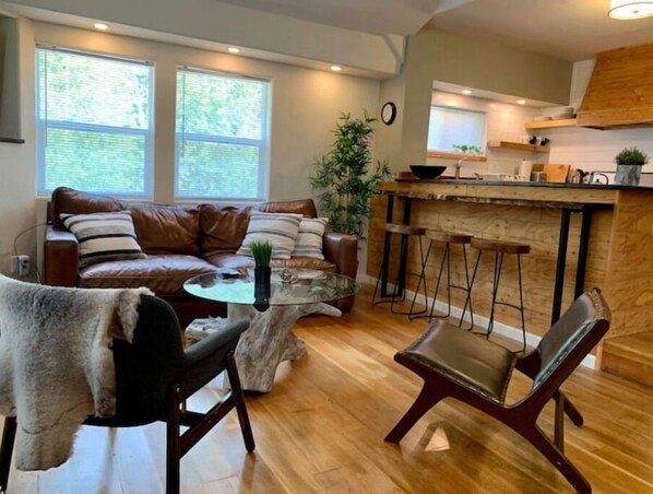A modern living room with a brown leather sofa, a glass coffee table, and a wooden dining area with bar stools.