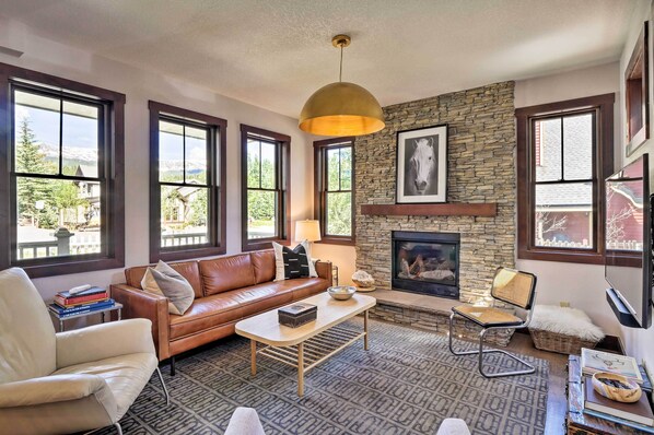 A cozy living room with a stone fireplace, leather sofa, and large windows offering a view of the outdoors.