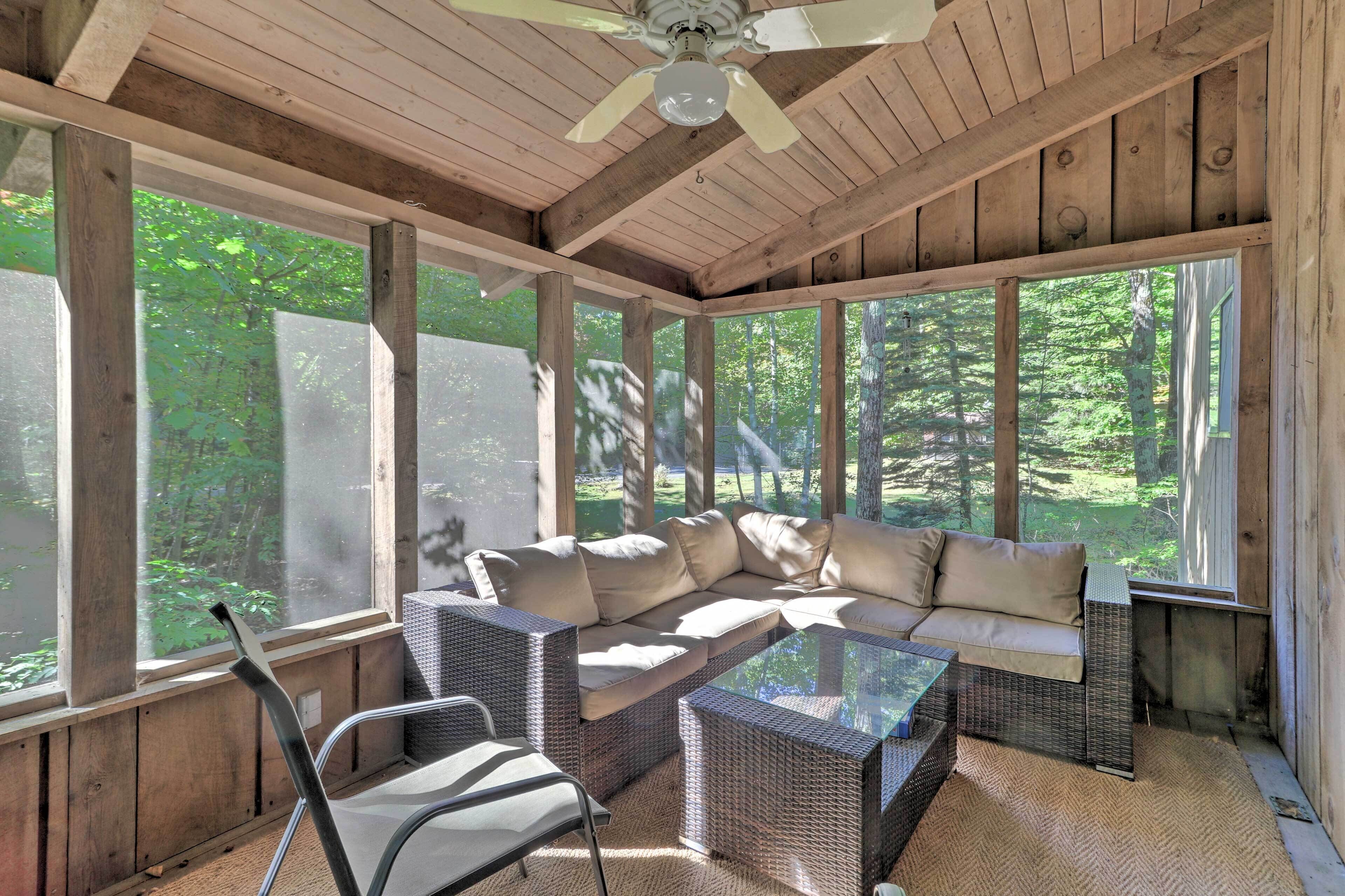 A covered outdoor seating area with a sofa and chairs, large windows, and a ceiling fan.
