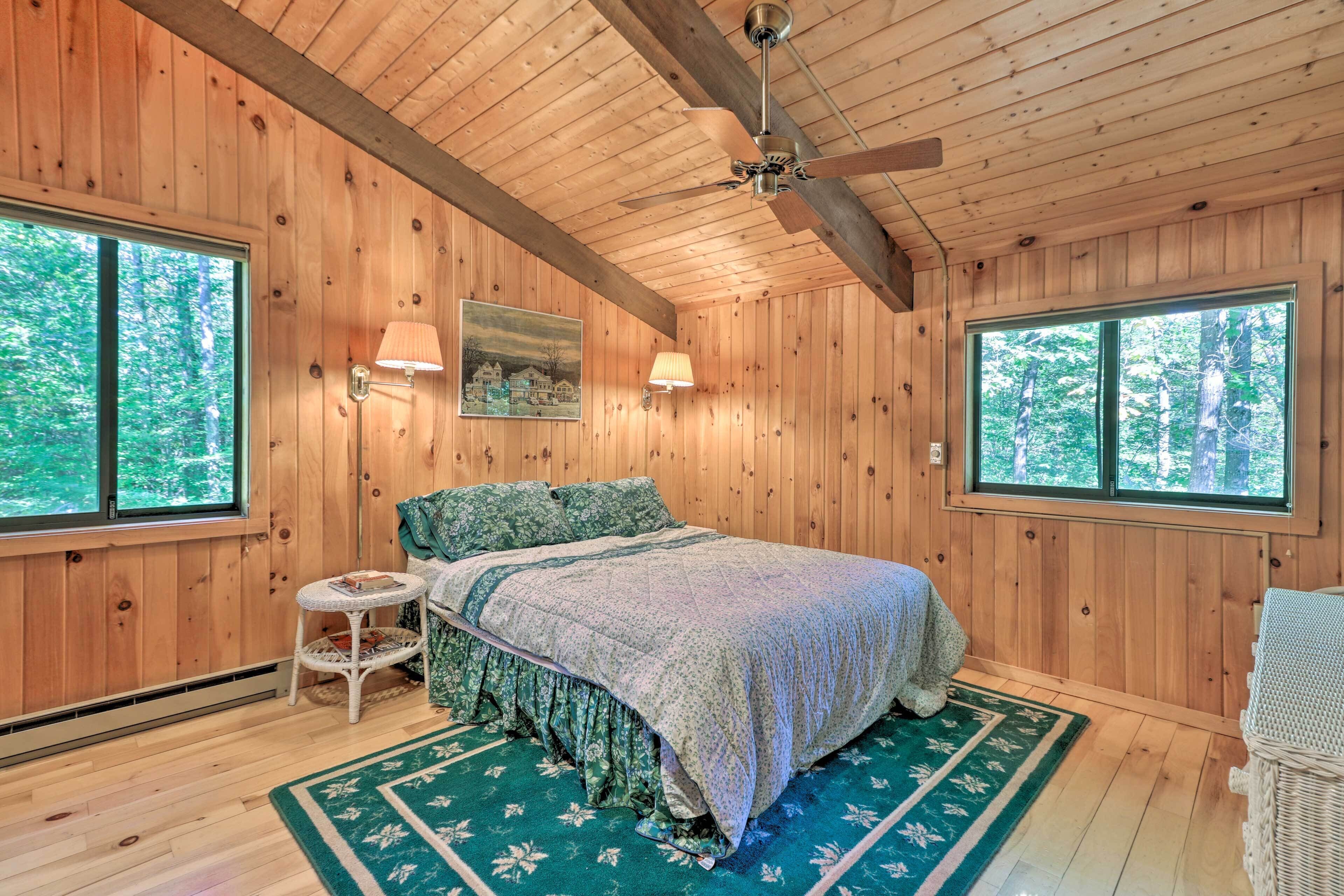 Une chambre aménagée dans une cabane en bois, avec un lit, deux lampes fixées au mur, un ventilateur de plafond et de grandes fenêtres.