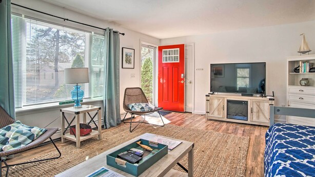 A well-lit room with a bed, a TV, a chair, a table with a lamp, and a red door. - Cape Cod Escape ~ 2 Mi To Beaches & Dining! (South Yarmouth)