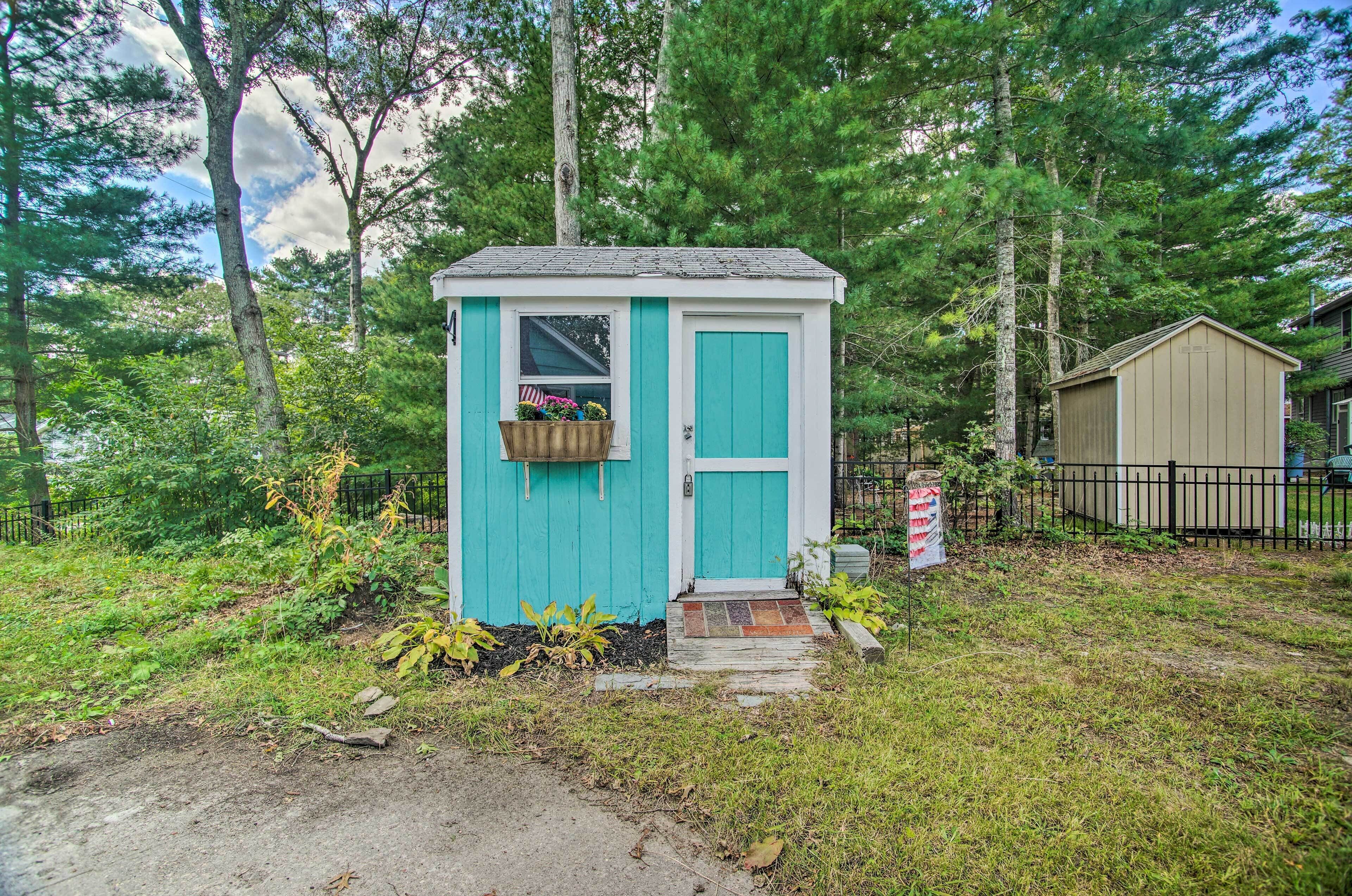 A small, single-story shed with a turquoise door and window, surrounded by a grassy area and trees.