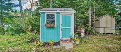 A small, single-story shed with a turquoise door and window, surrounded by a grassy area and trees.