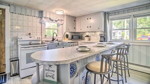 A kitchen with white cabinets, a large island with bar stools, a microwave, and a coffee maker. - Coastal Comfort Near the Bay! 'buttermilk Cottage' (Buzzards Bay)