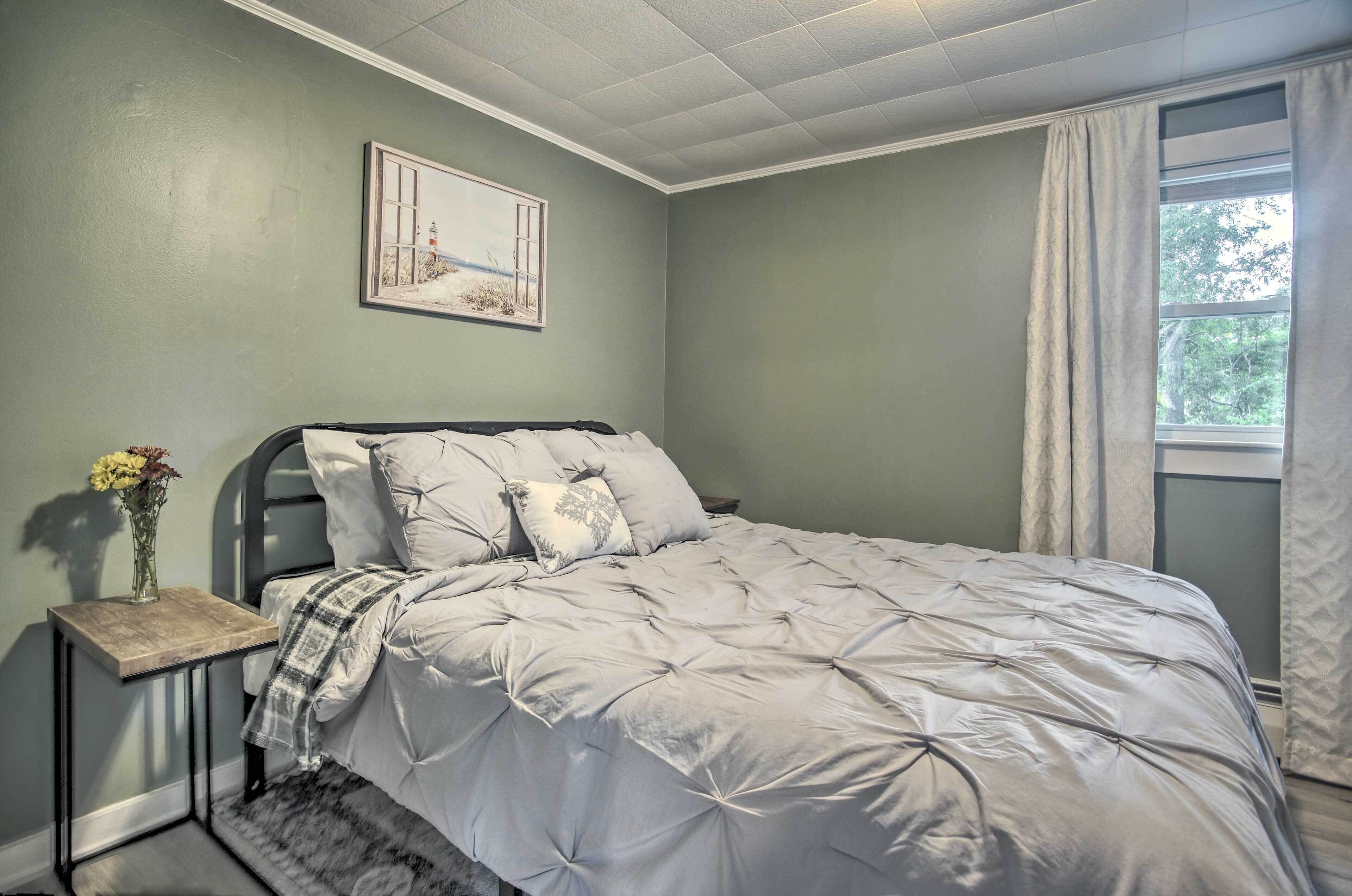 Un dormitorio con una cama, una mesita de noche, un cuadro enmarcado y una ventana con cortinas.